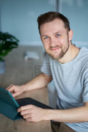 Téléchargez les photos : Attrayant barbu caucasien mâle freelance ingénieur logiciel, développeur ou web designer assis sur le lieu de travail dans le bureau à peine travailler à l'aide d'un ordinateur regardant la caméra souriant. Image de haute qualité - en image libre de droit