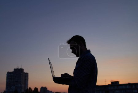 Téléchargez les photos : Développeur freelance ou l'homme il ingénieur travaillant en plein air en utilisant un ordinateur portable en silhouette au coucher du soleil avec des bâtiments. Adorable crépuscule en arrière-plan. Difficile de travailler le soir. Image de haute qualité - en image libre de droit