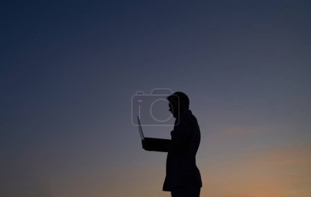 Téléchargez les photos : Homme développeur pigiste ou il ingénieur travaillant en plein air en utilisant un ordinateur portable en silhouette au coucher du soleil. Difficile de travailler le soir. Adorable crépuscule en arrière-plan. Image de haute qualité - en image libre de droit