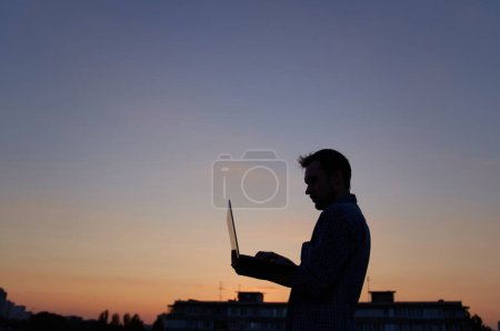 Téléchargez les photos : Homme ingénieur informatique ou pigiste en silhouette travaillant en plein air à l'aide d'un ordinateur portable. Coucher de soleil. Adorable crépuscule en arrière-plan. Difficile de travailler le soir. Image de haute qualité - en image libre de droit