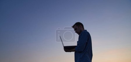Téléchargez les photos : Hardly working evening: male freelancer developer or it support manager in silhouette working outdoor using laptop at sunset. Adorable dusk at background. High quality image - en image libre de droit