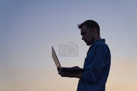 Téléchargez les photos : Hardly working evening concept. Adorable developer freelancer or it male engineer standing working outdoor using laptop in silhouette at sunset. Beautiful dusk at background. High quality image - en image libre de droit