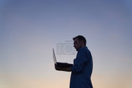 Téléchargez les photos : IT engineer man or freelancer in silhouette working outdoor using laptop. Sunset time. Adorable dusk at background. Hardly working evening. High quality image - en image libre de droit