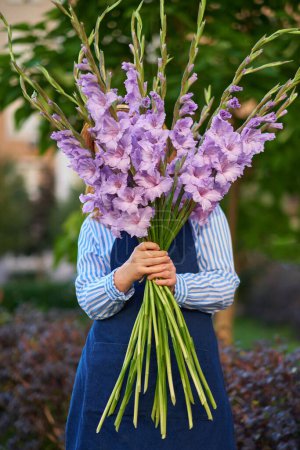 Photo for Red haired woman in blue shirt and hat holding bouquet of purple gladioluses outdoor, summer concept. No face seen. High quality photo - Royalty Free Image