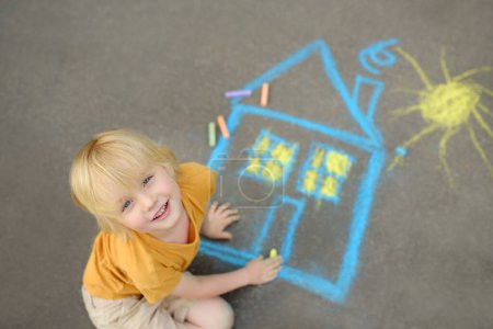 Little child boy is drawing house and sun painted with colored chalk on asphalt of sidewalk. Kids creative picture on gray background of road. Concepts of home and peaceful life all over the world.