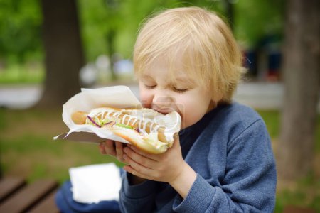 Little boy eating hot dog in public park. Child enjoying his to go meal outside. Fast food is a junk food. Overweight problem kids.