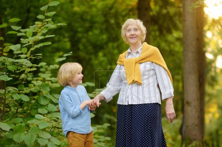 Cute grandson holding hands with his joyful elderly grandmother during walking at summer park. Two generations of family spend time together. Quality family time