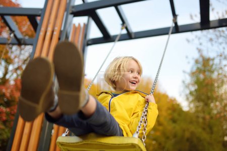 Téléchargez les photos : Petit garçon s'amusant sur une balançoire sur l'aire de jeux dans le parc public le jour de l'automne. Un enfant heureux aime se balancer. Loisirs actifs en plein air pour enfant en ville - en image libre de droit