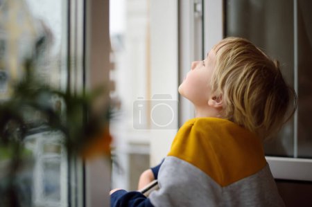 Foto de Little boy watching the rain outside at opened window. A child of primary school age is at risk of falling from a window from a great height. Dangers in the home for a small kid - Imagen libre de derechos