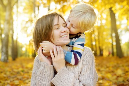 Foto de El niño abraza a su joven madre y la besa durante un paseo por el soleado parque de otoño. Amor y emociones entre mamá y el niño - Imagen libre de derechos