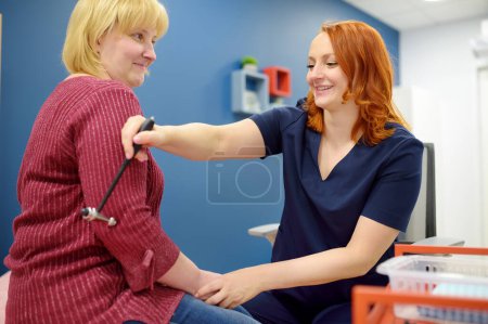Mature woman are at an appointment with a neurologist in modern clinic. Doctor is checking the neurological reflexes of patient with hammer. Neurology professional doctor at work