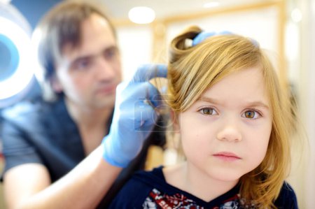 Portrait of cute little girl during appointment of dermatologist in modern clinic. Doctor examines child hair and scalp for lice and nits. Pediculosis and parasitic diseases are common in kids groups