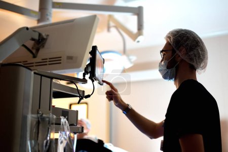 Ein Assistenzarzt überwacht den Fortschritt einer chirurgischen Operation auf einem tragbaren Bildschirm im Operationssaal eines modernen medizinischen Krankenhauses. Überwachung des Zustandes des Patienten während der Operation.