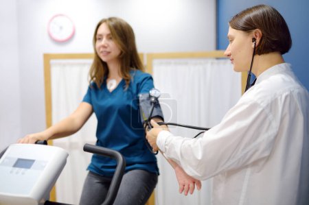 Female doctor cardiologist measures pressure with patient during bicycle exercise for examination cardiovascular system at medical clinic. Young woman training on bike simulator. Exercise bike