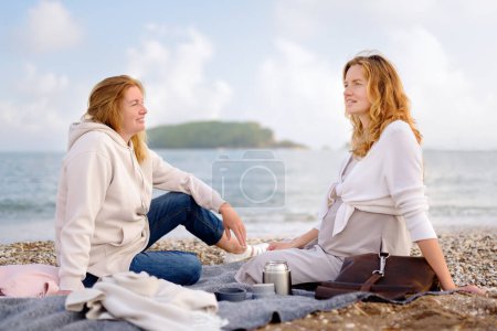 A redhead senior mother and her adult pregnant daughter are having a picnic by the sea shore. Happy meeting of a mother and her grown-up child. Family relationships between adult children and parents