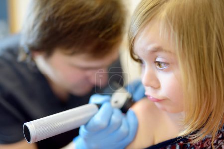 Foto de Un médico cuidadoso revisa los lunares en la piel de un niño pequeño. Un dermatólogo observa una erupción en la espalda de una niña usando un dermatoscopio. Bebé con mamá en una cita con un pediatra - Imagen libre de derechos