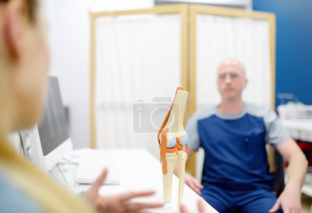 Foto de Ortopedista muestra al paciente un modelo anatómico de articulación de rodilla durante la consulta en clínica moderna. El médico explica las características de la estructura y los problemas de tratamiento de enfermedades. Primer plano de la articulación de la rodilla - Imagen libre de derechos
