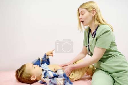 Lindo niño pequeño está en una cita con el pediatra. Médico gastroenterólogo palpando el vientre de un niño pequeño paciente acostado en el sofá en el consultorio médico. Enfermedades gastroenterológicas de los niños