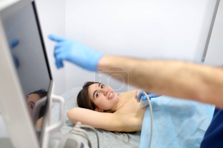 Säugetier untersucht Brüste und Lymphknoten einer Frau mit Ultraschall. Der Arzt zeigt dem Patienten einen Scan der Brustdrüsen auf dem Bildschirm. Brustkrebs-Bewusstsein