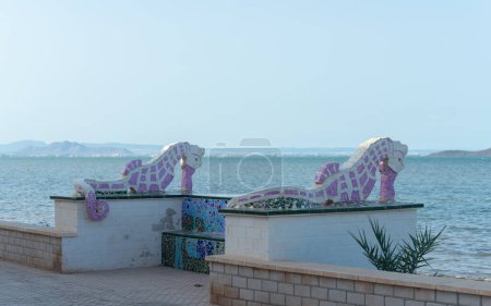 Foto de LOS NIETOS, ESPAÑA - 19 DE SEPTIEMBRE DE 2022 Esculturas de caballitos de mar en la orilla de la laguna Mar Menor, con la localidad de La Manga en el horizonte, Murcia, España - Imagen libre de derechos