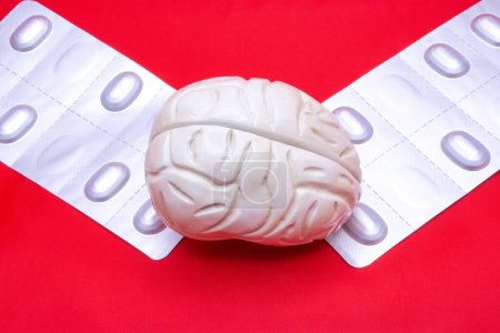 Die anatomische Figur des menschlichen Gehirns auf rotem Hintergrund, zusammen mit zwei Tablettenblasen für Kopfschmerzen oder nootrope Behandlung. Foto für den Einsatz in der Neurologie, Psychiatrie oder Inneren Medizin