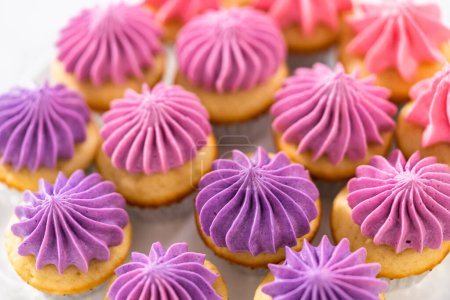 Téléchargez les photos : Mini cupcakes à la vanille fraîchement cuits avec glaçage beurre rose ombre sur un stand de gâteau blanc. - en image libre de droit