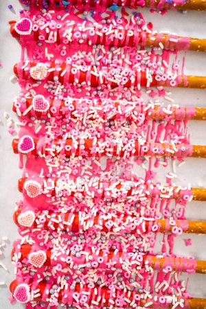 Drizzling melted chocolate over chocolate-dipped pretzels rods and decorating with sprinkles to make chocolate-covered pretzel rods for Valentines Day.