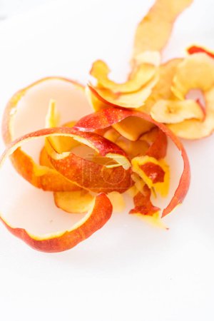 Téléchargez les photos : Peler la peau des pommes rouges avec un éplucheur de pommes de terre sur une planche à découper blanche. - en image libre de droit