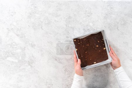 Téléchargez les photos : Pose plate. Poêle à gâteau au fromage carrée garnie de papier sulfurisé avec mélange de caramel pour préparer du caramel macadamia au chocolat. - en image libre de droit