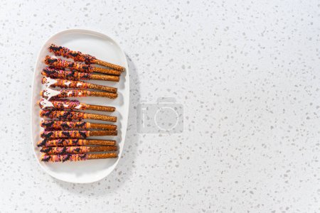 Photo for Flat lay. Halloween chocolate-covered pretzel rods with sprinkles on a white serving plate. - Royalty Free Image