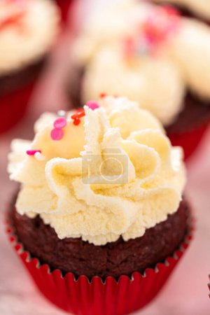 Foto de Piping glaseado de ganache de chocolate blanco en la parte superior de cupcakes de terciopelo rojo y cubierta con aspersiones. - Imagen libre de derechos
