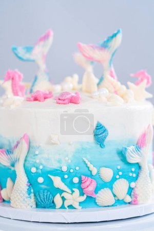 Téléchargez les photos : Gâteau à la vanille 3 couches sur le thème de la sirène décoré de queues de sirène au chocolat et de coquillages sur un stand de gâteau blanc. - en image libre de droit