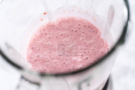 Blending ingredients for healthy breakfast strawberry banana smoothie in a kitchen blender.