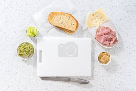 Téléchargez les photos : Pose plate. Assemblage de sandwichs au jambon, concombre et germe sur la planche à découper blanche. - en image libre de droit