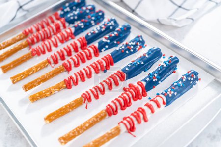 Foto de Barras de pretzel cubiertas de chocolate caseras decoradas como la bandera estadounidense secándose en una bandeja para hornear forrada con papel pergamino. - Imagen libre de derechos
