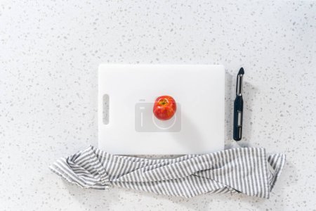 Téléchargez les photos : Flat lay. Peeling skin from red apples with a potato peeler on a white cutting board. - en image libre de droit