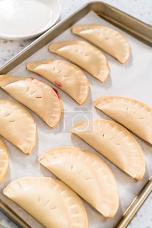 Téléchargez les photos : Empanadas de cerises douces non cuites sur une plaque à pâtisserie tapissée d'une plaque à pâtisserie en papier parchemin. - en image libre de droit