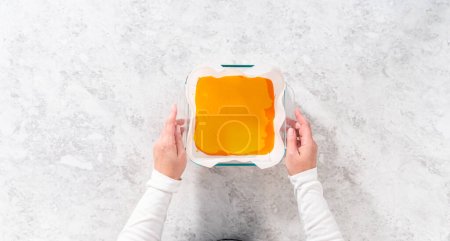Photo for Flat lay. Pouring fudge mixture into the square glass baking pan lined with parchment paper to prepare candy corn fudge. - Royalty Free Image
