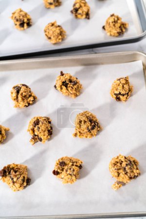 Téléchargez les photos : Ramasser la pâte à biscuits avec une cuillère à pâtisserie dans une plaque à pâtisserie doublée de papier parchemin pour faire cuire des biscuits aux noix de raisin à l'avoine molle. - en image libre de droit