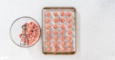 Flat lay. Scooping ground meat with dough scoop into a baking sheet lined with parchment paper to prepare oven-baked meatballs.