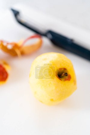 Téléchargez les photos : Peler la peau des pommes rouges avec un éplucheur de pommes de terre sur une planche à découper blanche. - en image libre de droit