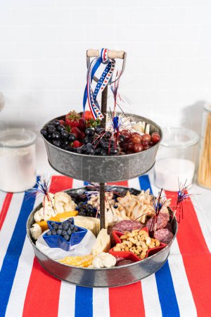 Foto de 4 de julio tablero de charcutería en un soporte de metal de dos niveles para servir lleno de queso, galletas, salami y frutas frescas - Imagen libre de derechos