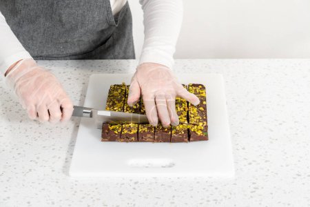 Téléchargez les photos : Cutting chocolate pistachio fudge with a large kitchen knife into square pieces on a white cutting board. - en image libre de droit