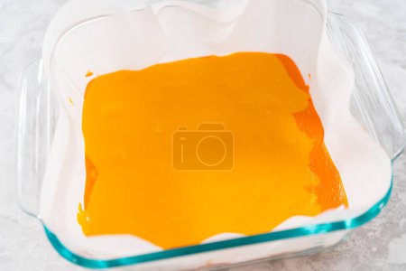 Photo for Pouring fudge mixture into the square glass baking pan lined with parchment paper to prepare candy corn fudge. - Royalty Free Image
