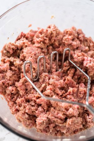 Photo for Mixing ingredients with a potatoe masher in a glass mixing bowl to prepare oven-baked meatballs. - Royalty Free Image