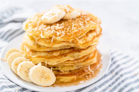 Photo for Stack of freshly baked coconut banana pancakes garnished with sliced bananas, toasted coconut, and maple syrup. - Royalty Free Image