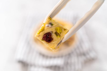 Photo for Holding homemade cranberry pistachio fudge square piece with kitchen tongs. - Royalty Free Image