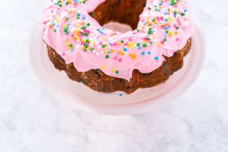 Téléchargez les photos : Gâteau bundt Funfettti givré à la crème au beurre à la vanille rose et décoré de saupoudres arc-en-ciel. - en image libre de droit