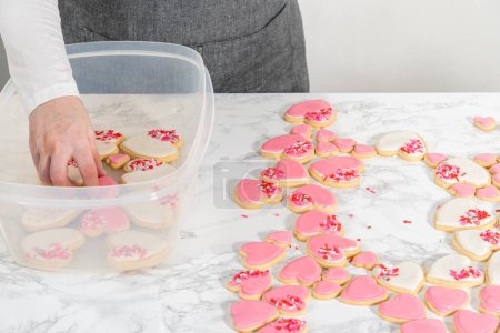 Almacenamiento de galletas de azúcar en forma de corazón con glaseado real rosa y blanco en un recipiente de plástico grande.