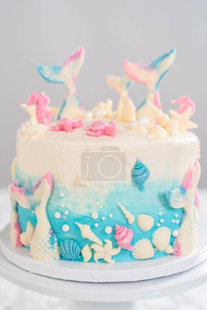 Photo for Mermaid-themed 3 layer vanilla cake decorated with chocolate mermaid tails and seashells on a white cake stand. - Royalty Free Image